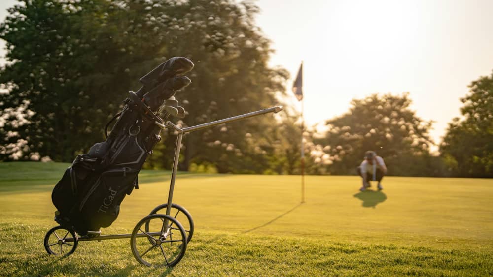 A sleek golf push cart bathed in golden sunlight on a peaceful golf course, with a golfer in the background lining up a putt near the flag.